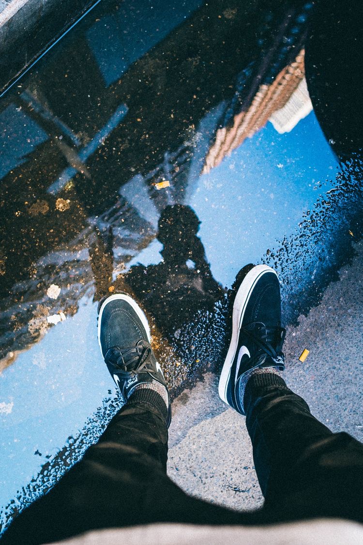 Point of view shot of skate shoes beside a street puddle reflecting the city