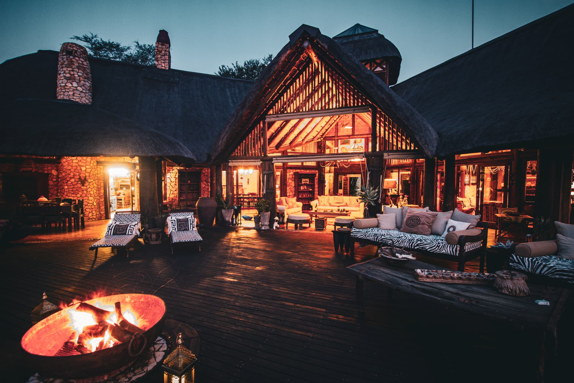 Thatched lodge deck at dusk with fire pit and warm interior glow